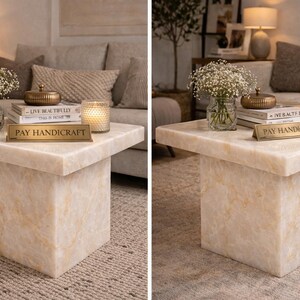 Puede incluir: Una mesa de centro cuadrada de m&aacute;rmol color crema con base de bloque. La mesa est&aacute; adornada con un jarr&oacute;n de flores blancas, libros, una vela y una caja dorada decorativa. Un letrero dorado dice "PAY HANDICRAFT".