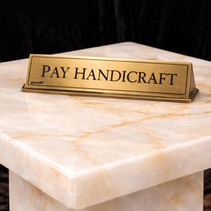 Puede incluir: Un letrero rectangular de lat&oacute;n con las palabras "PAY HANDICRAFT" en letras negras descansa sobre una superficie similar al m&aacute;rmol. El letrero est&aacute; inclinado y la superficie tiene un patr&oacute;n veteado crema y dorado. El fondo es oscuro.