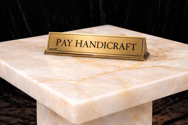 Puede incluir: Un letrero rectangular de lat&oacute;n con las palabras "PAY HANDICRAFT" en letras negras descansa sobre una superficie similar al m&aacute;rmol. El letrero est&aacute; inclinado y la superficie tiene un patr&oacute;n veteado crema y dorado. El fondo es oscuro.