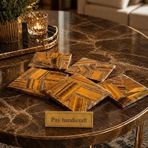 Könnte beinhalten: Sechs quadratische Untersetzer aus Tigeraugenstein liegen auf einem braunen Marmortisch. Die Untersetzer haben ein Mosaikmuster in Gold- und Brauntönen. Ein kleines Messingschild trägt die Aufschrift "Pay handicraft".