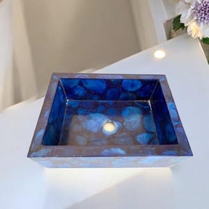 Könnte beinhalten: Ein quadratisches, blau-braunes dekoratives Waschbecken mit glänzendem Finish. Der Innenraum weist ein Muster aus blauen und braunen Formen auf. Das Waschbecken hat ein Abflussloch in der Mitte und steht auf einer weißen Oberfläche.