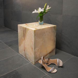 Könnte beinhalten: Ein quadratischer Beistelltisch aus weißem Onyx mit einer hellbraunen und grünen Marmorierung. Eine kleine Glasvase mit weißen Blumen steht auf dem Tisch. Zwei weiße Sandalen mit silbernen Riemen und Absatz stehen auf dem Boden neben dem Tisch.