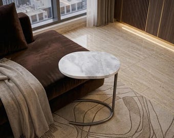 May include: A C-shaped side table featuring a round white marble top with grey veining, supported by a curved dark metal base. The table is positioned next to a brown sofa draped with a light grey throw blanket on a patterned area rug.