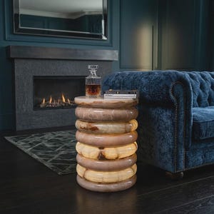May include: A cylindrical side table crafted from stacked, polished stone rings with a warm, translucent, beige and brown marbled pattern. The table features an internal light source that illuminates the stone, topped with a glass decanter.