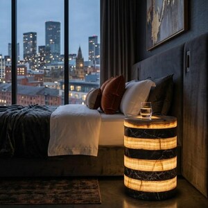 May include: A cylindrical bedside table made of alternating bands of dark black marble and translucent, glowing amber-colored stone. The table features a smooth, polished finish and emits a warm, soft light from its illuminated stone sections.