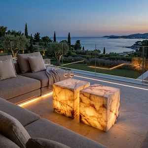 May include: Two illuminated cube-shaped side tables made of translucent white and amber veined stone. The tables feature a warm internal glow and are positioned on a stone patio next to a gray fabric sofa with matching throw pillows.