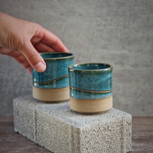 Könnte beinhalten: Zwei handgefertigte Keramikbecher mit türkisfarbener Glasur und beigem Boden. Die Becher sind zylindrisch und stehen auf einem grauen Betonblock. Die Becher werden auf einer Holzoberfläche präsentiert.