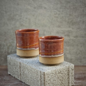May include: Two ceramic cups with a reddish-brown glaze and a tan base. The cups are displayed on a grey concrete block, highlighting their unique design. These handmade cups are ideal for hot beverages, such as tea or coffee.