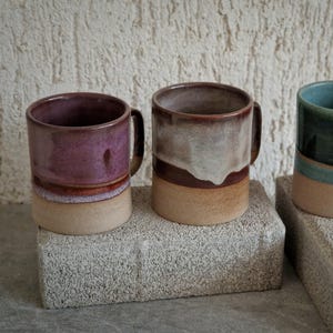 Op de afbeelding: Drie keramische mokken met handvatten, elk met een uniek kleurenschema. De mokken zijn in tinten paars, bruin en groen, met een beige basis. Ze staan op grijze rechthoekige blokken tegen een witte achtergrond.