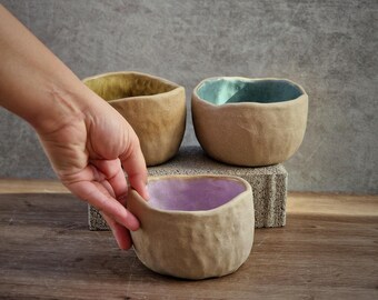 May include: Three handmade ceramic bowls with textured exteriors and colorful interiors. One bowl has a purple interior, another has a teal interior, and the third has a yellow interior. The bowls are displayed on a wooden surface.