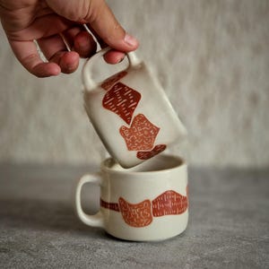 May include: Two white ceramic mugs with brown abstract designs. One mug is held above the other, both with a handle. The mugs are on a grey surface. The mugs have a cream-coloured background with brown abstract shapes.