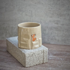 Peut inclure: Une tasse en céramique beige avec une surface texturée et une petite illustration de renard orange. La tasse est posée sur un bloc rectangulaire gris, avec un fond en bois.