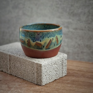 May include: A handcrafted ceramic bowl with a distinctive glaze. The bowl has a dark red base, blending into a yellow and blue-green centre, and a dark blue rim. It sits on a light grey brick on a wooden surface.