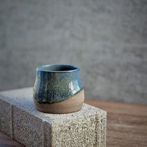May include: A handcrafted ceramic cup with a rounded form. The upper half is glazed in a mottled blue, fading to a natural tan base. The cup rests on a textured, light grey brick, with a wooden surface in the background.