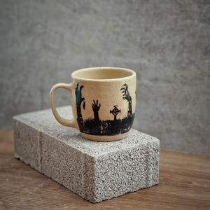 May include: A beige ceramic mug with a black and blue zombie design. The design shows zombie hands reaching up from a graveyard with a cross. The mug has a handle and is on a grey brick.