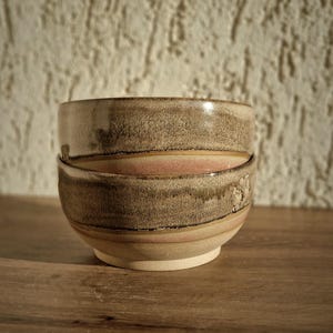 以下が含まれることがあります： 手作りの陶器のボウルが2つ重ねられています。ボウルは、茶色、タン、ピンクの色合いが混ざり合った、質感のあるアースカラーです。質感のあるオフホワイトの壁と木製の表面に置かれています。