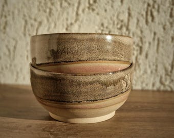 May include: Two handmade ceramic bowls stacked on top of each other. The bowls have a textured, earthy appearance with a mix of brown, tan, and pink hues. The bowls are set against a textured, off-white wall and a wooden surface.