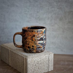 May include: A ceramic mug with a textured surface and a brown, orange, and blue glaze. The mug has a handle and is displayed on a grey concrete block. The mug is suitable for coffee, tea, or other beverages.