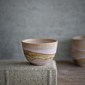 May include: A ceramic bowl with a pink and brown glaze. The bowl is placed on a grey concrete block. The bowl has a rounded shape and a smooth surface. The background is a blurred grey.