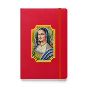 Könnte beinhalten: Ein rotes Notizbuch mit einem elastischen Bandverschluss. Das Cover zeigt ein gerahmtes Porträt einer Frau in traditioneller Kleidung, mit blauem Kopftuch und aufwendigem Schmuck. Das Porträt ist vor einem floralen Hintergrund platziert.
