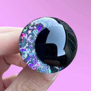 A hand painted glitter safety eye with off-set black pupils and colorful glitter with black stars and moons.