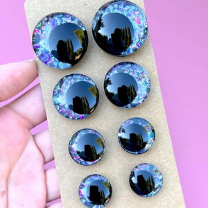 Four pairs of hand painted glitter safety eyes with off-set black pupils and colorful glitter with black stars and moons. They are arranged in two rows on a brown card.
