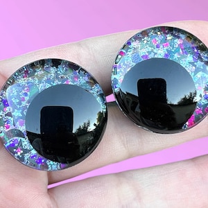 A pair of hand-painted glitter safety eyes with off-set black pupils and colorful glitter with black stars and moons.