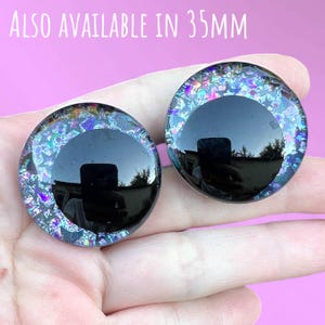 A pair of hand-painted glitter safety eyes with off-set black pupils and colorful glitter with black stars and moons. The text also available in 35mm is written above the eyes.