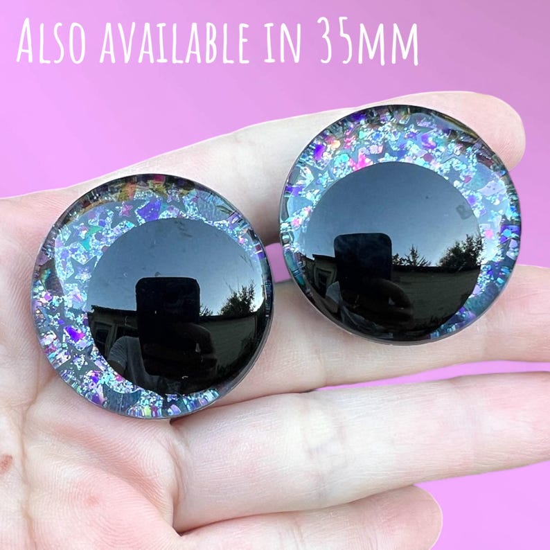 A pair of hand-painted glitter safety eyes with off-set black pupils and colorful glitter with black stars and moons. The text also available in 35mm is written above the eyes.