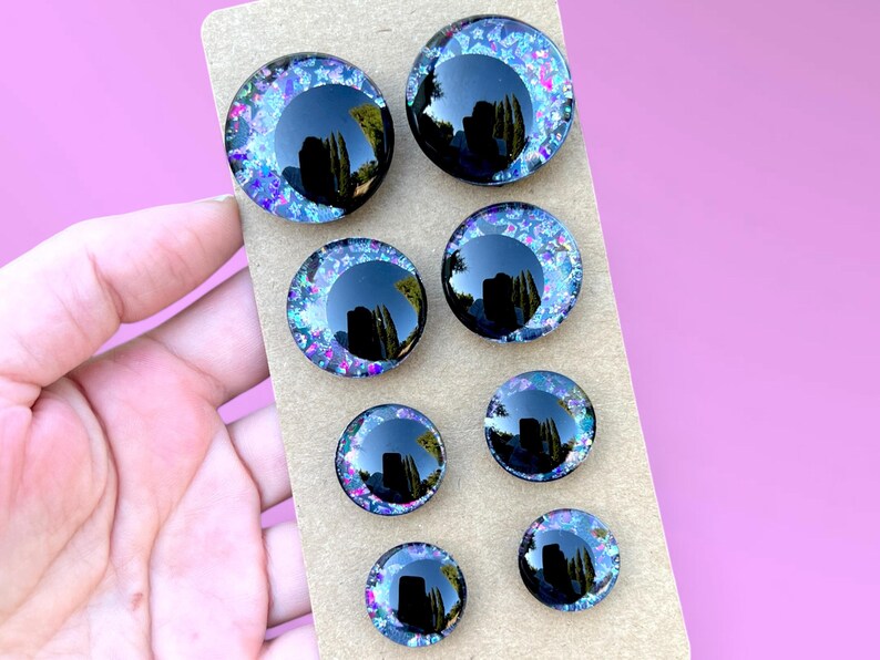 Four pairs of hand painted glitter safety eyes with off-set black pupils and colorful glitter with black stars and moons. They are arranged in two rows on a brown card.