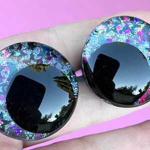 An upclose photo of a pair of hand-painted glitter safety eyes with off-set black pupils and colorful glitter with black stars and moons.