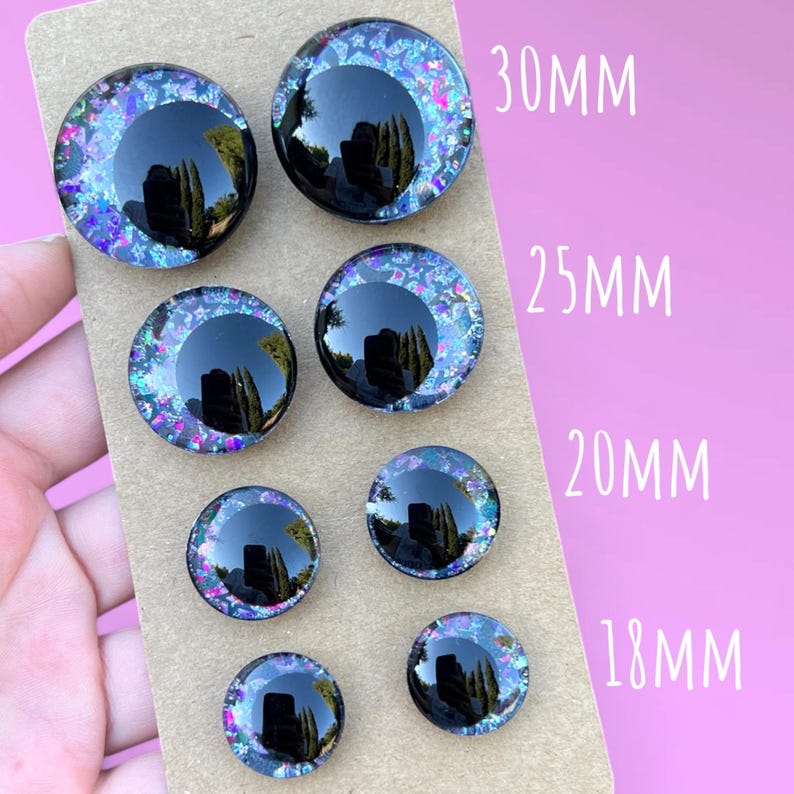 Four pairs of hand painted glitter safety eyes with off-set black pupils and colorful glitter with black stars and moons. They are arranged in two rows on a brown card. Each pair has a size written to the right of them, 30mm, 25mm, 20mm, and 18mm.