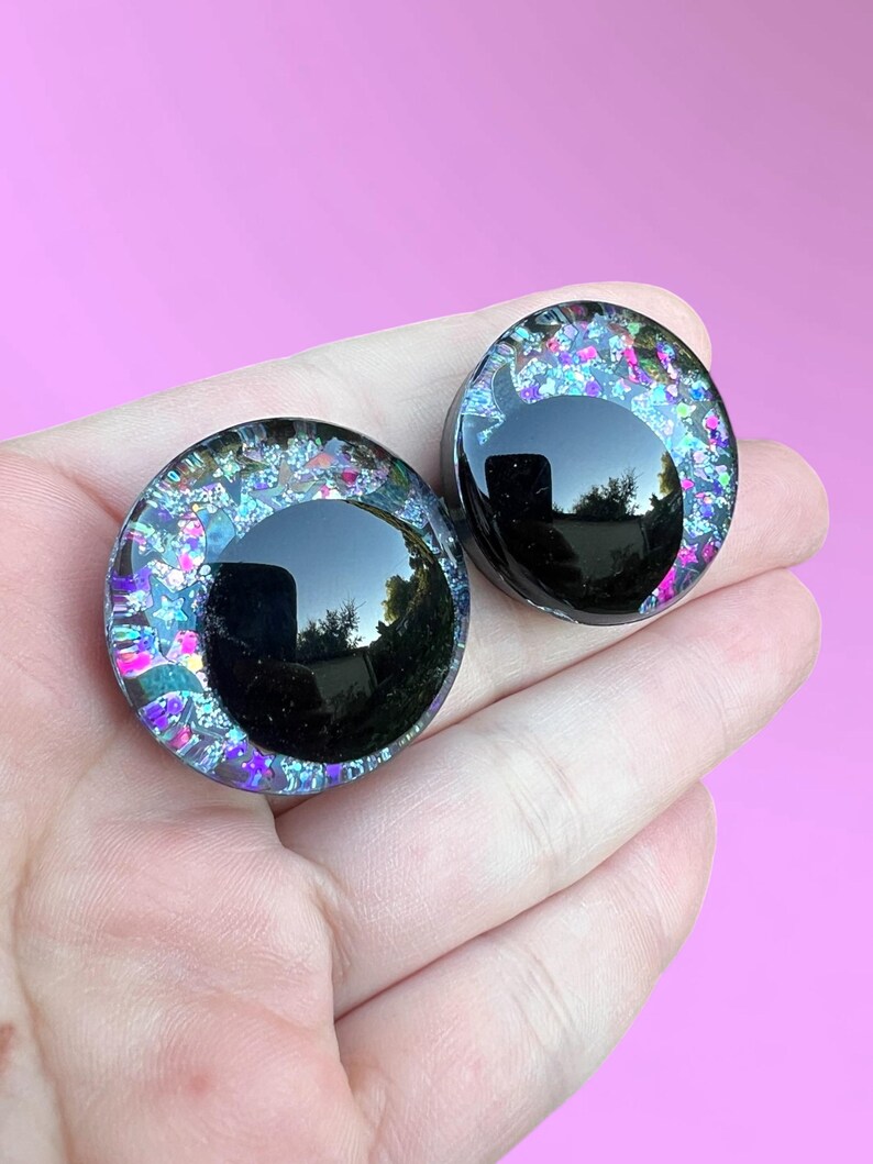 A pair of hand-painted glitter safety eyes with off-set black pupils and colorful glitter with black stars and moons.