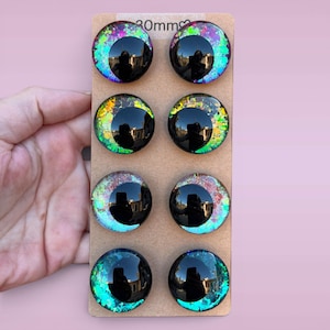 May include: Eight circular cabochons arranged in four pairs on a rectangular cardboard backing labeled 30mm. Each piece features a large black pupil set against a vibrant, multi-colored iridescent background with flecks of green, blue, and purple.