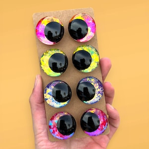 Könnte beinhalten: Acht kreisförmige Cabochons, angeordnet in vier Paaren auf einer rechteckigen Pappunterlage. Jedes Stück zeigt eine große, glänzend schwarze Pupille vor einem lebendigen, irisierenden Hintergrund mit rosa, gelben, blauen und violetten Folien.