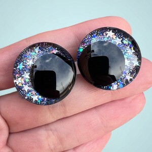 A pair of hand painted plastic safety eyes with off-set black pupils with silver glitter stars and galaxy colored glitter.