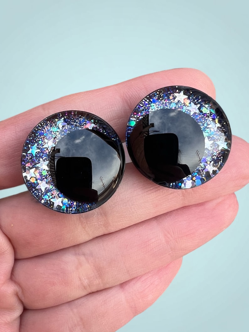 A pair of hand painted plastic safety eyes with off-set black pupils with silver glitter stars and galaxy colored glitter.