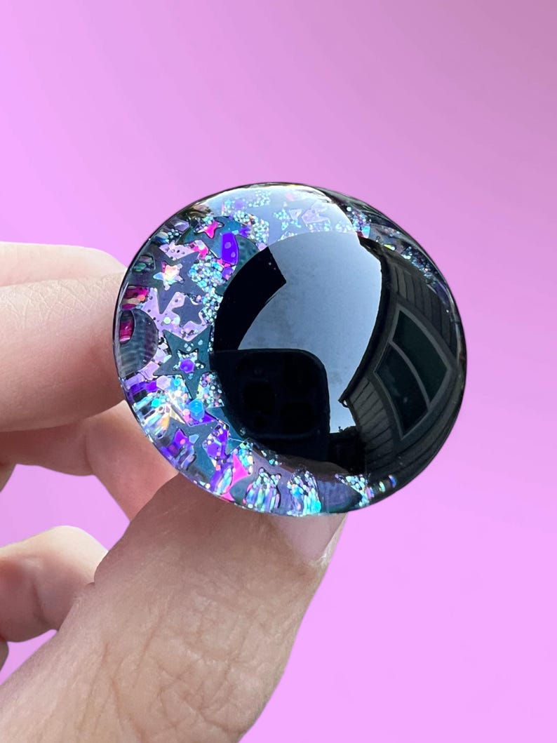 A hand painted glitter safety eye with off-set black pupils and colorful glitter with black stars and moons.