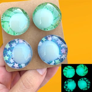 Puede incluir: Cuatro cabujones circulares con una cúpula central azul pálido lisa. Los dos superiores muestran un patrón de placa de circuito verde, mientras que los dos inferiores tienen un fondo de purpurina azul con formas de estrellas y lunas. Un recuadro muestra su brillo.
