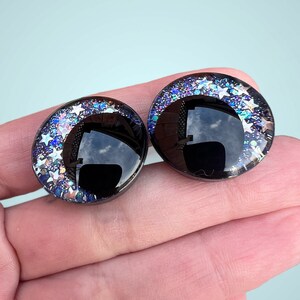 A pair of hand painted plastic safety eyes with off-set black pupils with silver glitter stars and galaxy colored glitter.