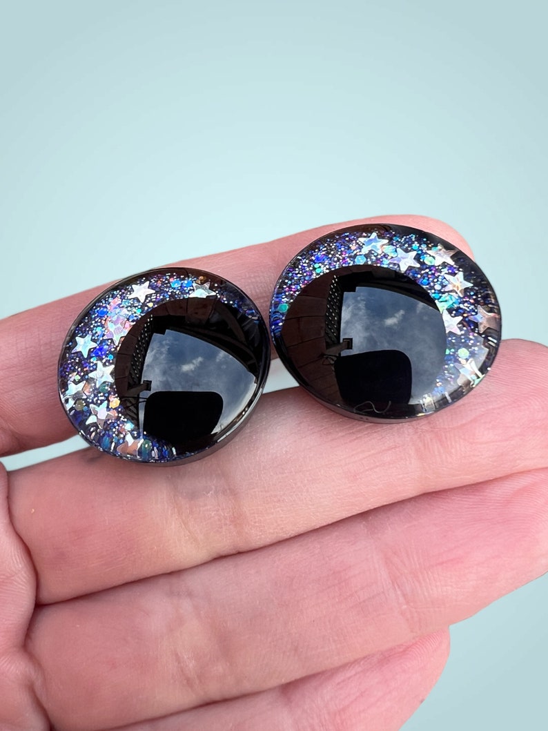A pair of hand painted plastic safety eyes with off-set black pupils with silver glitter stars and galaxy colored glitter.