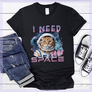 Puede incluir: Camiseta negra con un gato atigrado con un casco de astronauta, sobre un fondo cósmico. El texto "I NEED SOME SPACE" está impreso encima y debajo de la ilustración del gato. La camiseta se combina con vaqueros azules y zapatillas de lona negras.