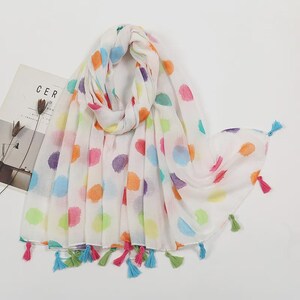 May include: A white scarf with a colorful polka dot pattern in shades of orange, pink, purple, green, and blue. The scarf has tassels in matching colors at the ends. The scarf is made of a lightweight fabric.