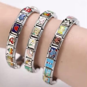 Op de afbeelding: Drie zilveren bedelarmbanden met kleurrijke emaille ontwerpen. Elke armband heeft rechthoekige bedels met verschillende afbeeldingen, waaronder harten, bloemen en een raket. De armbanden worden om een pols gedragen.