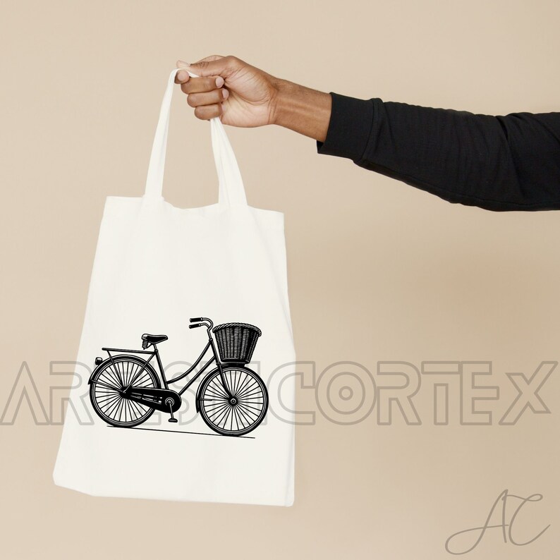 Puede incluir: Una bolsa de tela blanca con una ilustraci&oacute;n en blanco y negro de una bicicleta con una cesta en la parte delantera. La bolsa tiene un asa negra y est&aacute; siendo sostenida por una mano.