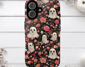 Puede incluir: Una funda de teléfono con un fondo oscuro cubierto de flores rosas y fantasmas blancos de dibujos animados. La funda tiene un acabado brillante y un recorte para la cámara. Los fantasmas tienen ojos redondos simples y bocas abiertas.