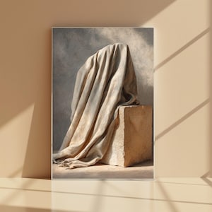 May include: A framed art print featuring a draped fabric over a stone block. The fabric is a light beige color, with soft folds and shadows. The stone block is a rough, textured beige. The background is a muted gray.
