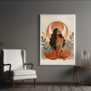 May include: A framed art print featuring a woman in a meditative pose. The artwork depicts a woman with long brown hair, wearing a light green top and orange pants, seated in front of a crescent moon and foliage. The color palette includes warm earth tones.