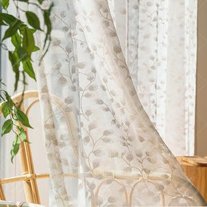 Puede incluir: Cortinas transparentes de color beige con un delicado estampado de hojas. Las cortinas son semitransparentes, permitiendo que la luz se filtre. El diseño de hojas es de un tono beige más claro, creando un contraste sutil. Las cortinas cuelgan frente a una ventana.