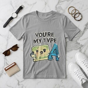May include: A gray t-shirt with the phrase "YOU'RE MY TYPE" and a cartoon graphic of a green square and a blue letter "A". The shirt is surrounded by accessories including a watch, sunglasses, and bracelets.